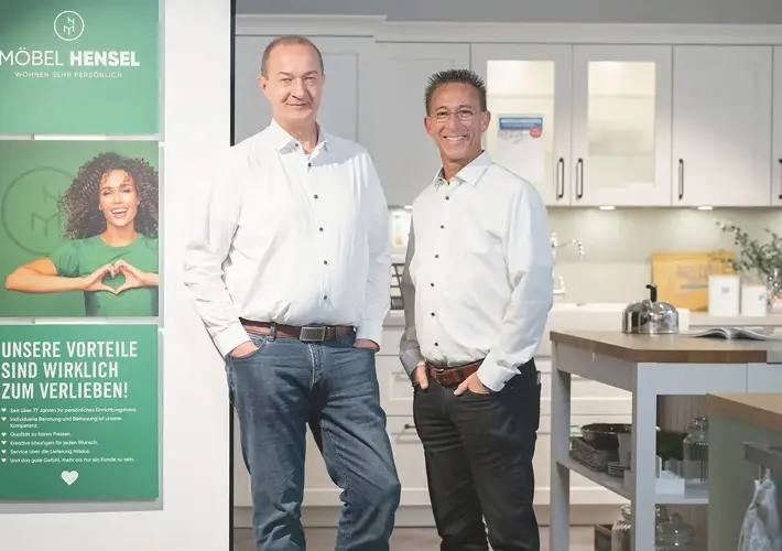 Zwei Männer vor weißer Einbauküche neben Plakat mit Text MÖBEL HENSEL und VORTEILE